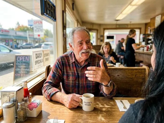 A man in his early seventies with a short white beard sitting at a diner table with a coffee mug, talking with his hands.